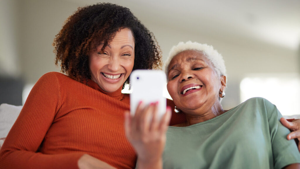 Close-up of a smiling younger woman with curly hair and an older woman with white hair looking at a white smartphone. Both are laughing and embracing, creating a heartwarming scene.