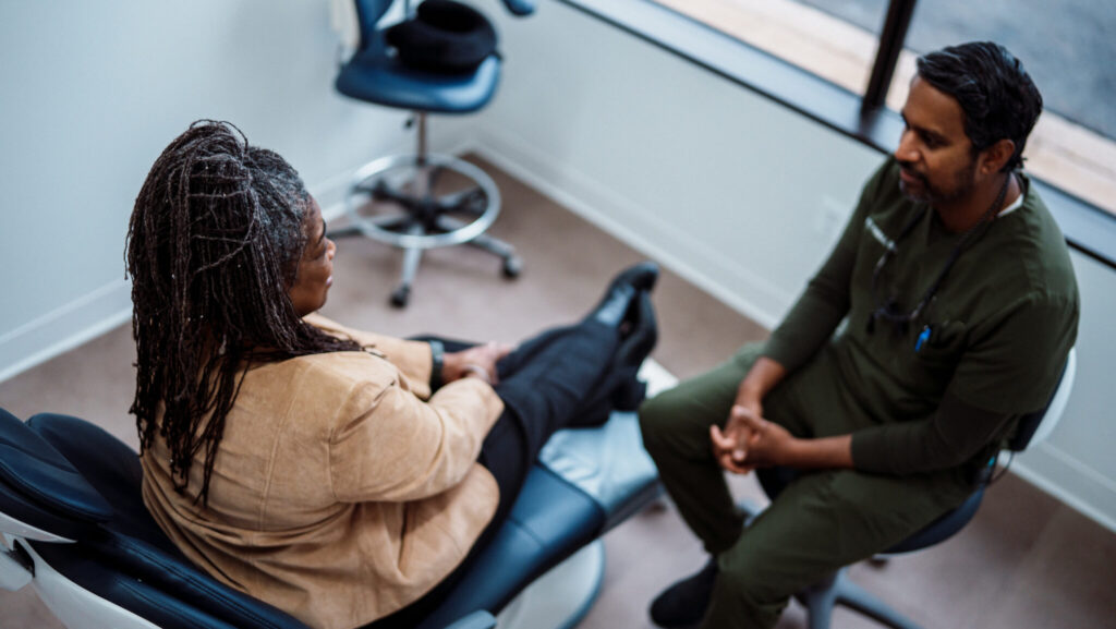 A male dentist of Indian descent wearing green medical scrubs sits on a stool and talks with a Black senior woman in a modern and bright dental examination room.