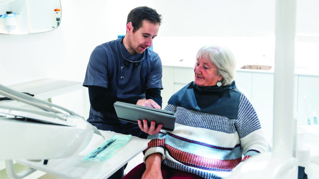 Dentist showing diagnosis and treatment plan on digital tablet to a senior woman patient during consultation in a modern dental clinic