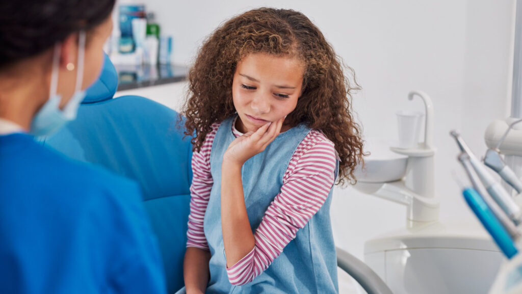 Girl holding cheek while sitting in dental chair showing sign on pain to female dentist. Child experiencing discomfort during clinic visit. Woman dentist observing young patient appears to be suffering from a severe toothache or jaw discomfort.