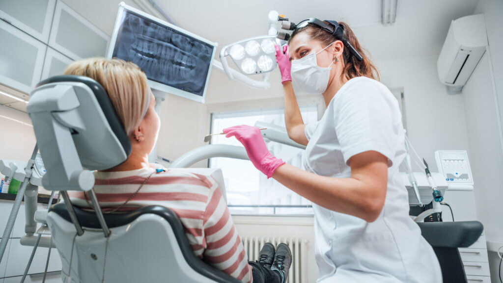 Dental clinic patient visit modern medical ward. Dentist female doctor dressed uniform in magnifying glasses pointing light. Patient sitting in chair. Health care and medicare industry concept image