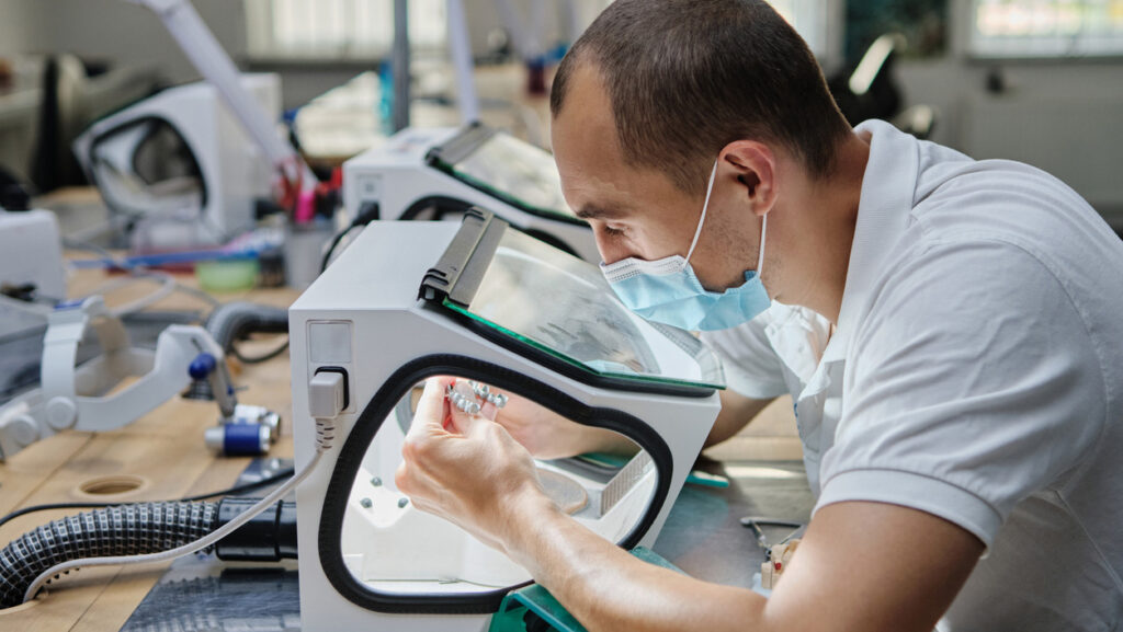 Dentist hand mills tooth crowns created on 3d printer for metal. Dental technician working with ceramic crowns in protective box at the laboratory. Metal processing